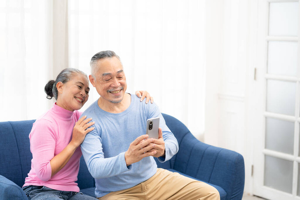 Feliz sonrisa asiática pareja mayor sentado en el sofá y el uso de teléfono inteligente para videollamada en línea en casa sala de estar. Sonriendo madura Video Call Conferencia charla chat concepto en línea. - Foto, imagen