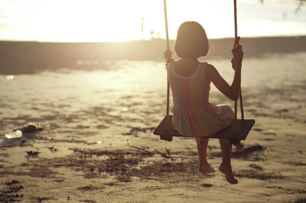 Achteraanzicht van eenzaam aziatisch meisje zit op de houten schommel met zonsondergang in de achtergrond - Foto, afbeelding