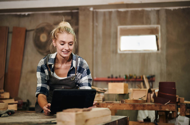 Femme blanche charpentier portant un tablier calcule et dessine des boiseries sur tablette numérique tout en travaillant sur son lieu de travail. - Photo, image