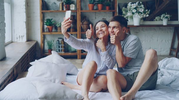 Funny young people boyfriend and girlfriend are taking selfie with silly faces having fun sitting on bed at home. Self-portrait and rerlationship concept. - Photo, image