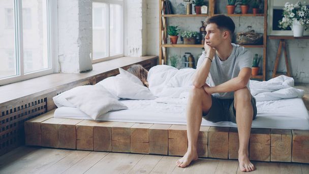 Sad man is sitting on bed after quarrel with his wife touching his face and looking at window while his wife is lying in bed with her back to him. Family crisis and people concept. - Foto, Bild