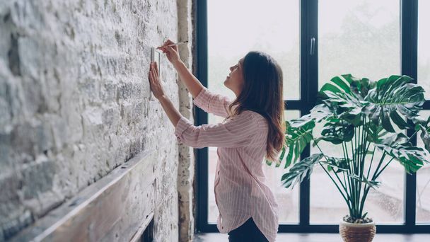 Creative young woman is decorating her loft style apartment choosing place on brick wall for beautiful picture and marking spot with pencil. Creativity and interior concept. - Photo, Image