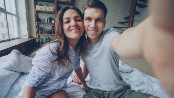 Point of view shot of loving couple taking selfie together posing having fun while sitting on bed at home. Nice modern interior and large windows are visible. - Photo, image