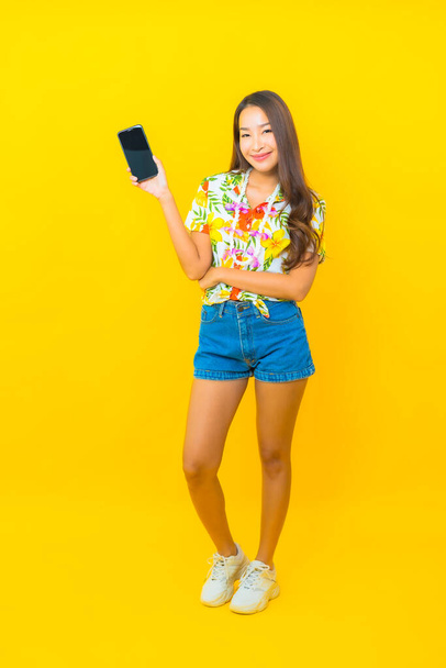 Porträt schöne junge asiatische Frau tragen buntes Hemd für sonkran Festival verwenden Smartphone auf gelbem Hintergrund - Foto, Bild