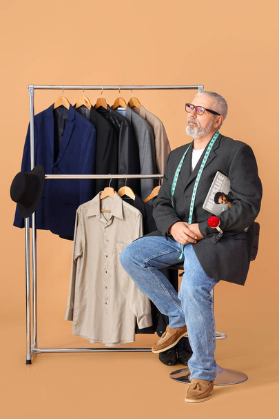 Diseñador de moda maduro con revista, estante y ropa sobre fondo beige - Foto, imagen