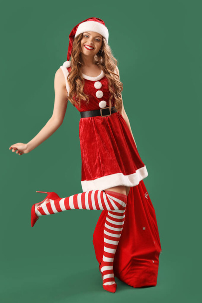 Pretty young woman in Santa costume with bag on green background - 写真・画像