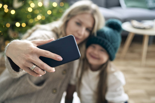 Caucásico madre e hija tomando selfie con teléfono inteligente con árbol de Navidad en el fondo  - Foto, Imagen