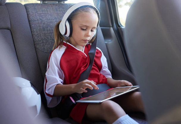 Child with tablet, headphones and safety belt in the car going home after soccer match, game or practice. Young girl in sports gear playing educational games online on tech device after training. - Foto, immagini