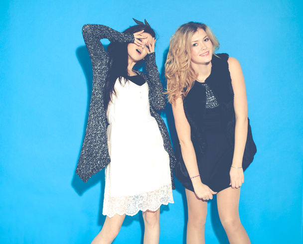 Fashionable beautiful young girlfriends standing together. Having funny and posing. Looking at camera. Indoor. Studio. Fashionable beautiful young girlfriends standing together near a blue background. - Foto, imagen