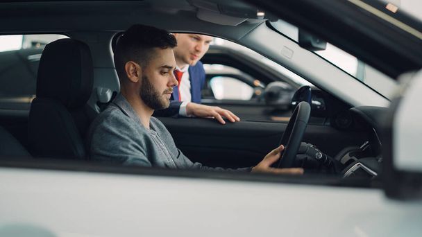 Confident man car buyer is sitting in drivers seat in new automobile inside modern showroom and talking to salesman standing near car window. Guy is holding steering wheel. - Foto, Imagen