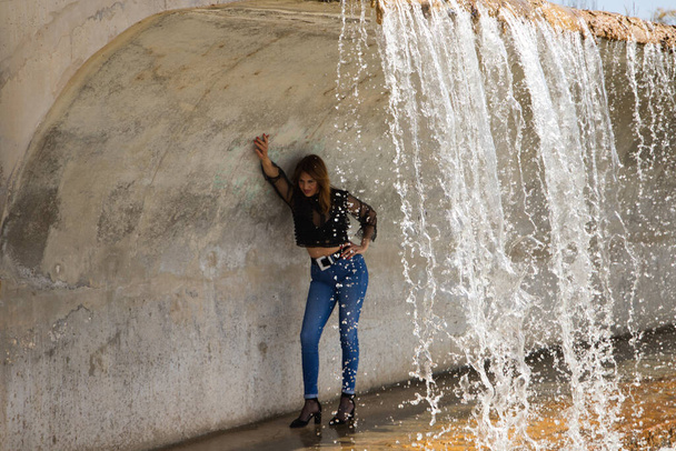 Красива, модно одягнена зріла жінка під великим водоспадом в парку. Жінка позує руками, витягнутими на стіну чуттєвим чином. Концепція моди та краси
. - Фото, зображення