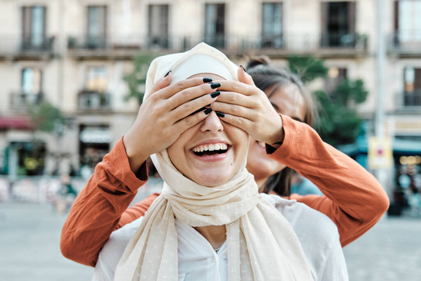 若い女性が友人の目を覆いながら通りの屋外で彼女を驚かせた。多民族友好の概念. - 写真・画像