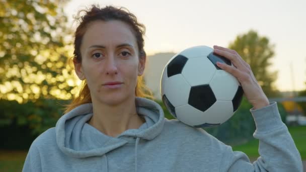 Kobieta trzymająca piłkę nożną patrzącą w obiektyw i uśmiechającą się w podświetleniu słońca. Portret piłkarki nożnej w parku o zachodzie słońca. Tło zachodu słońca. Wysokiej jakości materiał 4k - Materiał filmowy, wideo