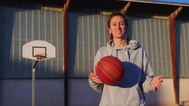 Vrouwensport concept. Een jonge vrolijke vrouw in sportkleding op een basketbalveld voor een wedstrijd. Oranje basketbal in handen. Hoge kwaliteit 4k beeldmateriaal - Video
