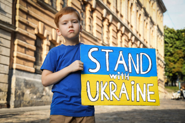 Sad boy holding poster in colors of national flag and words Stand with Ukraine on city street - Foto, afbeelding