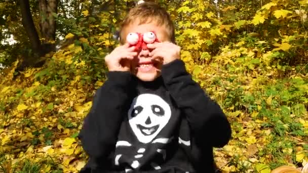 Un niño con un traje de esqueleto negro se apoya en sus aterradores ojos rojos y lo asusta durante Halloween. Hojas amarillas de otoño en el fondo - Imágenes, Vídeo