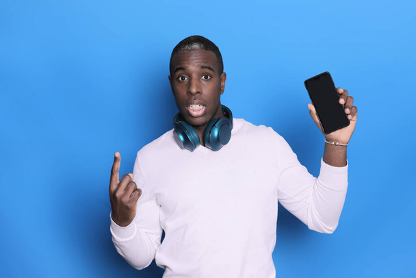 Emotional black man holds a smartphone in his hands, showing his finger up in a blank space. Blue background - Foto, afbeelding