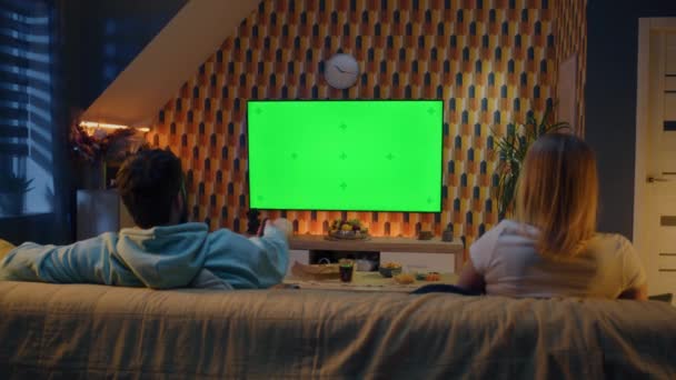 リビングルームでソファに座っているカップルは、軽食でテレビで速報ニュース、映画やシリーズを見ています。妻と夫は夜一緒に家で休んでいます。緑の画面だ。クロマキー。ホームシネマ. - 映像、動画
