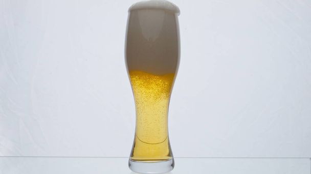 Helder glas vol schuimig smakelijk bier overlopen van de rand in super slow motion close-up. Licht borrelend schuim dat uit transparante beker op een witte ondergrond stroomt. Alcoholdrankconcept - Foto, afbeelding