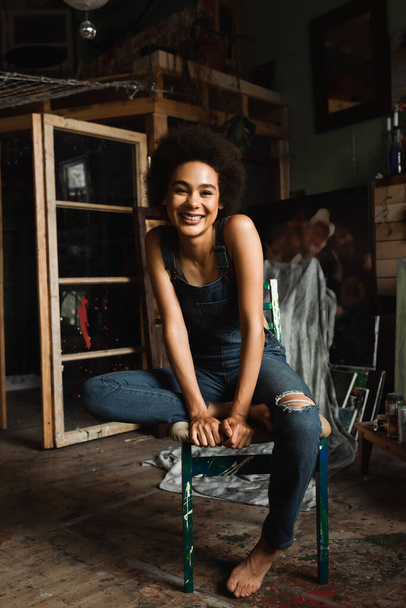 barefoot african american woman in overalls sitting in art studio and looking at camera - Foto, immagini