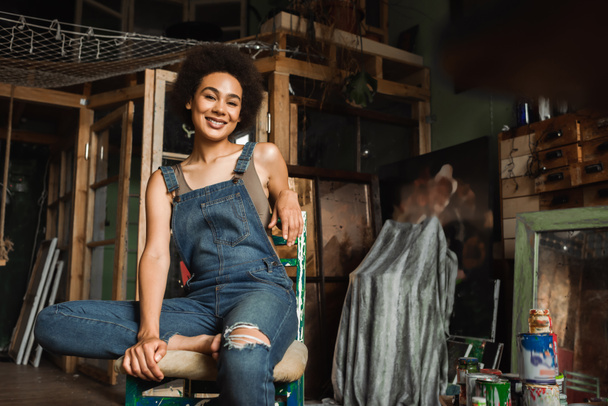 Αφροαμερικανή γυναίκα με denim φόρμες κάθεται στο στούντιο τέχνης και χαμογελά στην κάμερα - Φωτογραφία, εικόνα