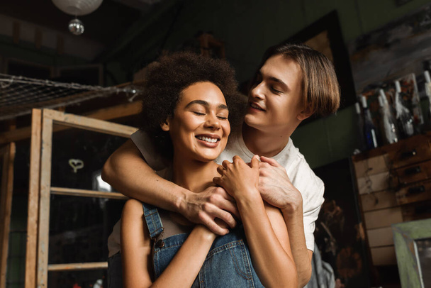 happy artist embracing pretty african american girlfriend smiling with closed eyes in workshop - Photo, Image