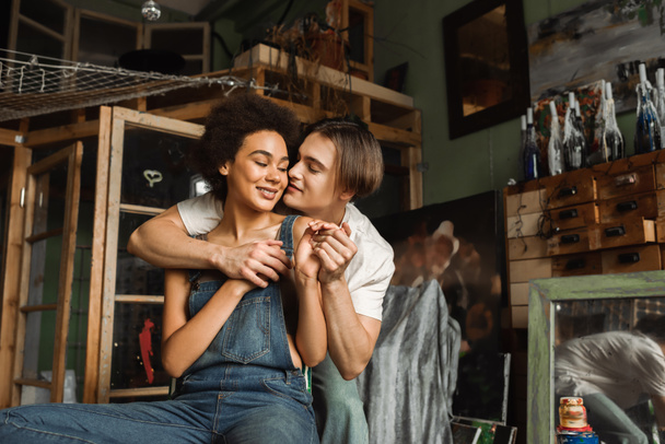 smiling african american woman in denim overalls holding hands with boyfriend in art studio - Photo, Image