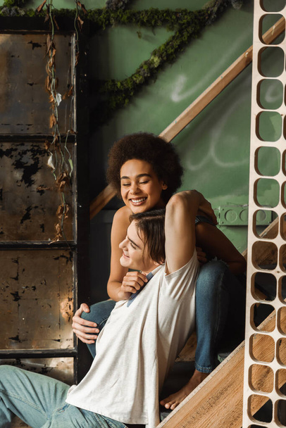 mulher americana africana alegre com olhos fechados abraçando com homem feliz enquanto sentado em escadas na oficina de arte - Foto, Imagem
