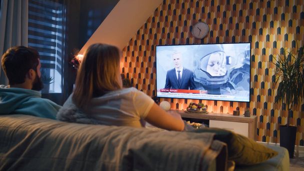 Paar zittend op de bank met snacks in de woonkamer, kijkend naar nieuws op tv over astronauten, ruimtemissie, het verkennen van de baan om de Aarde. Vrouw en man rusten en chillen thuis in het weekend 's nachts - Foto, afbeelding