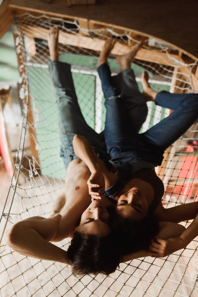top view of shirtless man in jeans and african american woman in top and overalls lying in mesh hammock above workshop - 写真・画像