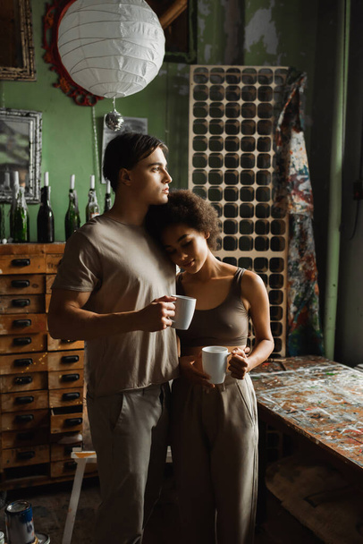 african american woman with cup of tea leaning on dreamy boyfriend looking away in workshop - Фото, изображение