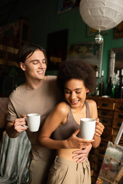 happy man with cup of tea hugging cheerful girlfriend in art workshop - Photo, Image