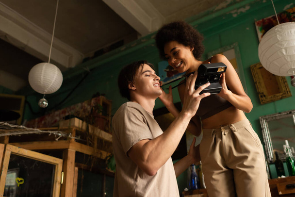 low angle view of smiling interracial artists holding vintage camera in art studio - Zdjęcie, obraz
