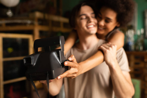 blurred african american woman taking selfie on vintage camera with happy boyfriend in workshop - Photo, Image