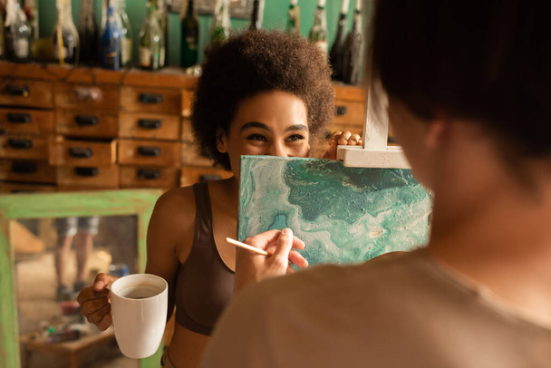 alegre afroamericana mujer con taza de té escondido detrás del caballete cerca borrosa artista - Foto, Imagen