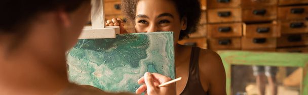 cheerful african american woman looking from behind easel on blurred boyfriend in workshop, banner - Photo, Image
