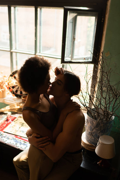 vista ad alto angolo di uomo muscoloso senza maglietta che abbraccia e bacia donna africana americana vicino alla finestra - Foto, immagini
