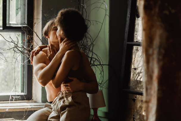apasionada pareja interracial abrazando y besándose cerca de plantas secas y ventana en taller de arte - Foto, Imagen