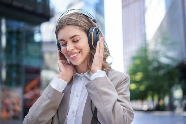 Ritratto di donna aziendale sorridente, ragazza in giacca e cravatta che ascolta musica in cuffia, seduta nel centro della città. - Foto, immagini