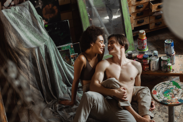 happy shirtless artist and african american woman in top sitting on floor near paints in art studio - Photo, Image