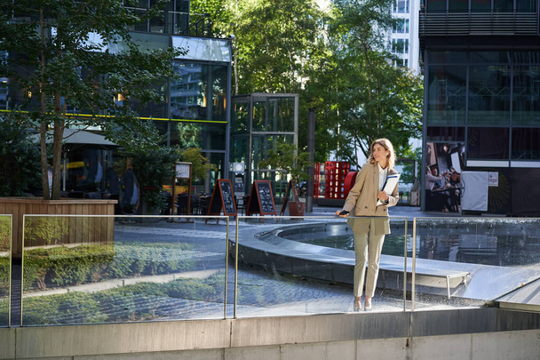 Корпоративна жінка в костюмі, що стоїть з упевненістю в бежевому костюмі з робочими документами на відкритому повітрі, позує в центрі міста
. - Фото, зображення