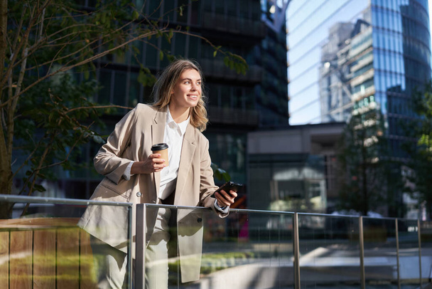 Succesvolle zakenvrouw in pak, drinkt haar koffie en gebruikt mobiele telefoon, staat op straat, heeft een lunchpauze. - Foto, afbeelding