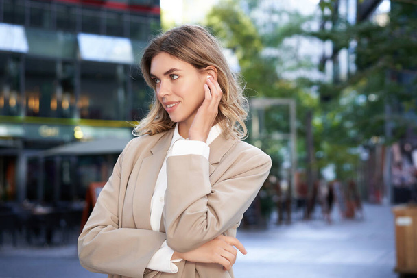 Nahaufnahme Porträt einer blonden Geschäftsfrau, selbstbewusste Geschäftsfrau mit Kopfhörern und beigem Anzug, die draußen auf der Straße im Stadtzentrum posiert. - Foto, Bild
