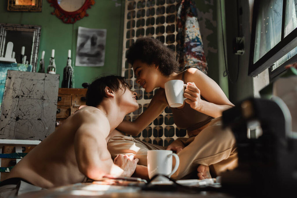 smiling african american woman sitting on windowsill with tea cup and looking at sexy shirtless boyfriend  - Φωτογραφία, εικόνα