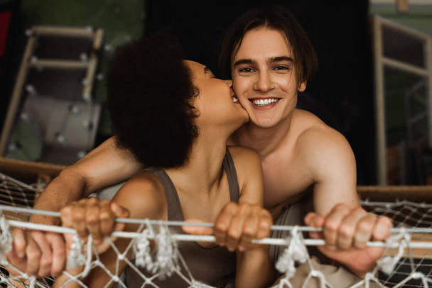 sexy african american woman kissing happy shirtless boyfriend looking at camera from hammock - Foto, imagen