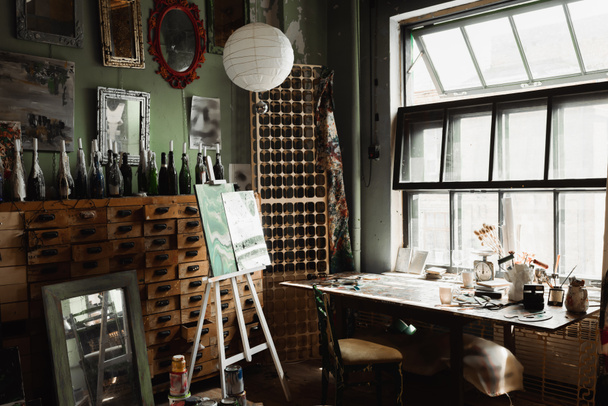 художественная мастерская с большим окном и старинной мебелью возле бутылок со свечами и зеркалами на стене  - Фото, изображение