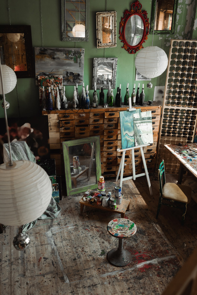 art studio with vintage furniture and mirrors with paintings on wall near easel and cans with paints - Foto, Imagem