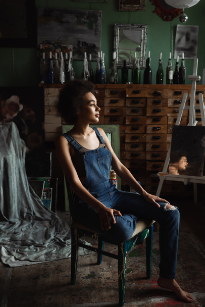 brunette african american woman in overalls sitting in workshop with vintage decor and looking away - Valokuva, kuva