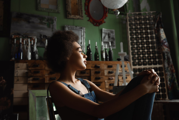 νέα και σκεπτική Αφροαμερικανή γυναίκα με denim φόρμες να κάθεται στο εργαστήρι τέχνης και να κοιτάζει αλλού - Φωτογραφία, εικόνα