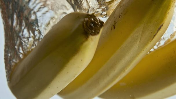 Großaufnahme Bananen fallen Wasser im hellen Hintergrund. Gelbe tropische Früchte geben transparente Flüssigkeit ab. Süße exotische Zutat zum Frühstück waschen. Schöne Lebensmittelwerbung. Gesunde Ernährung.  - Foto, Bild
